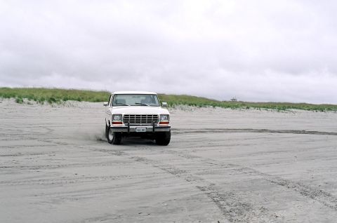 1979 Ford Bronco - A white 1979 Ford Bronco navigates a sandy terrain under an overcast sky.