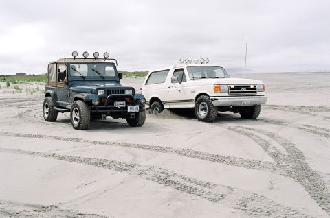 1979 Ford Bronco - Two off-road vehicles, a Jeep and a Ford Bronco, sit on a sandy beach under an overcast sky.