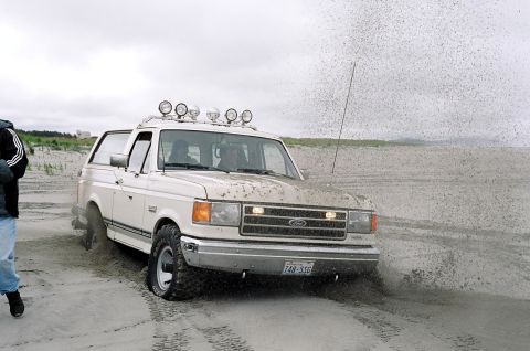 1979 Ford Bronco - A white Ford Bronco navigates a sandy terrain, with a person standing to the left and a flag pole in the background.