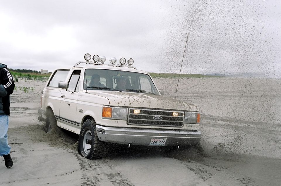 White Ford Bronco driving on sand, kicking up spray. Has roof lights and a license plate reading 748-SSG