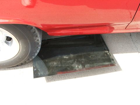 2005 Ford Mustang - A close-up view of a red car's side panel reveals a broken window resting on the ground beneath the vehicle.