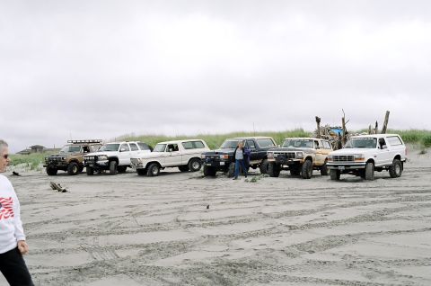 1979 Ford Bronco - A group of Ford Broncos are lined up on a sandy beach under an overcast sky.