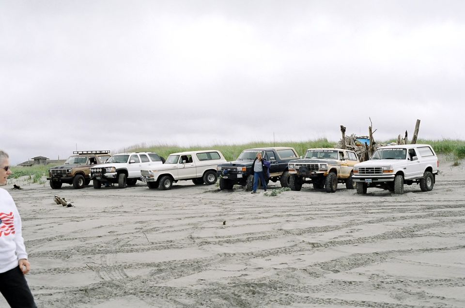 Several Ford Broncos parked on a sandy beach with dunes in the background