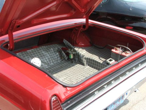 2005 Ford Mustang - Open trunk of a red classic car, revealing spare tire and battery. Another vehicle is seen in the background.
