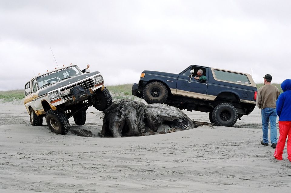 Two 1979 Ford Broncos off-roading on a beach, one climbing over driftwood