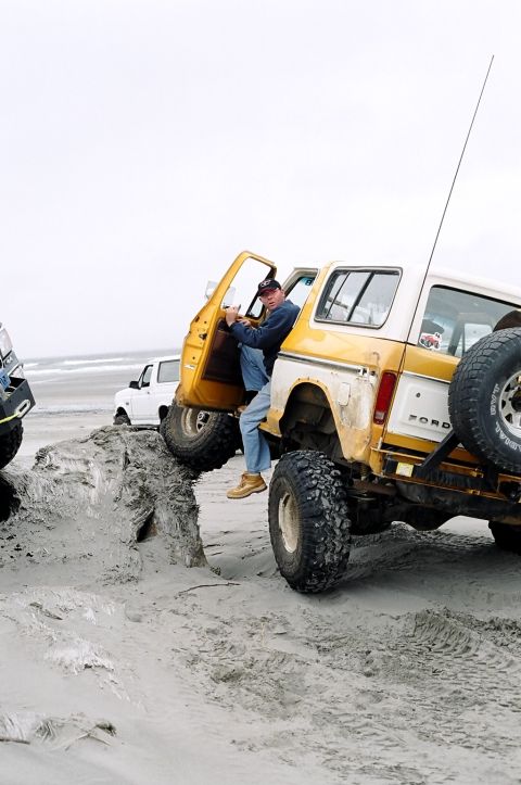 1979 Ford Bronco - A man exits a yellow Ford Bronco as it navigates a sandy incline on the beach, with another Bronco visible in the background.