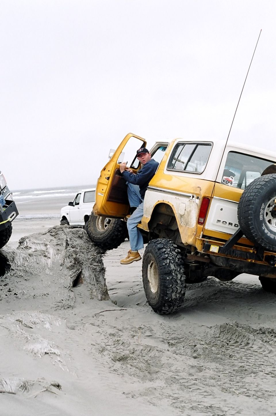 Guy climbing into yellow Ford Bronco on beach, another white Bronco behind it
