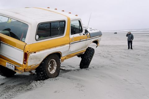 1979 Ford Bronco - A man stands on a beach near a yellow and white Ford Bronco partially buried in sand.