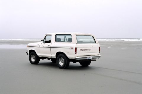 1979 Ford Bronco - A white 1979 Ford Bronco is seen on a beach, with the ocean and sky visible behind it.