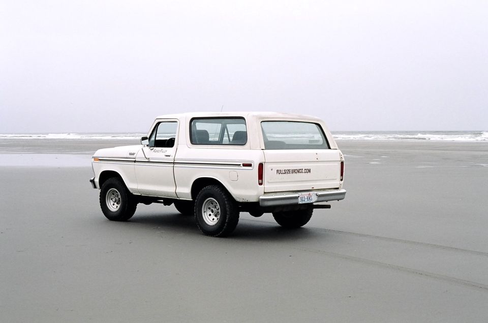 White 1979 Ford Bronco parked on a sandy beach with the ocean in the background