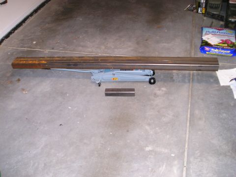 1996 Ford Bronco - A floor jack is positioned under two long metal beams, with a short metal piece on the concrete floor.
