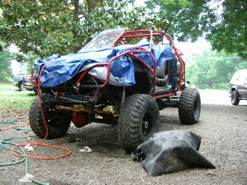 1985 Ford Bronco - An off-road vehicle with a custom roll cage and tarp cover sits on a gravel surface.