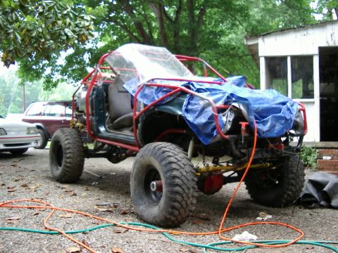 1985 Ford Bronco - This heavily modified off-road vehicle features a prominent roll cage and is partially covered by a blue tarp. Another vehicle is parked ne…