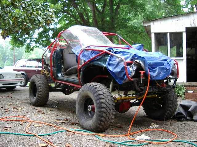 Off-road vehicle with roll cage, tarp, and large tires. Another vehicle visible