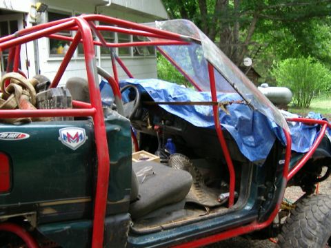 1985 Ford Bronco - Modified off-road vehicle with extensive roll cage and tarped interior, parked outdoors.