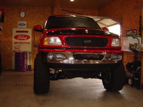 1978 Ford Bronco - Head-on front view of a red lifted Ford F-150 inside an OSB-walled shop, showing the chrome bumper, mesh grille with blue Ford oval, and an…
