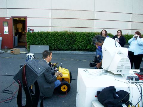 2000 Ford Excursion - A man in a suit drives a yellow ride-on toy car during what appears to be a film or TV production.
