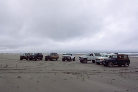 1996 Ford Bronco - Several SUVs and trucks are parked on a sandy beach near the ocean on an overcast day.