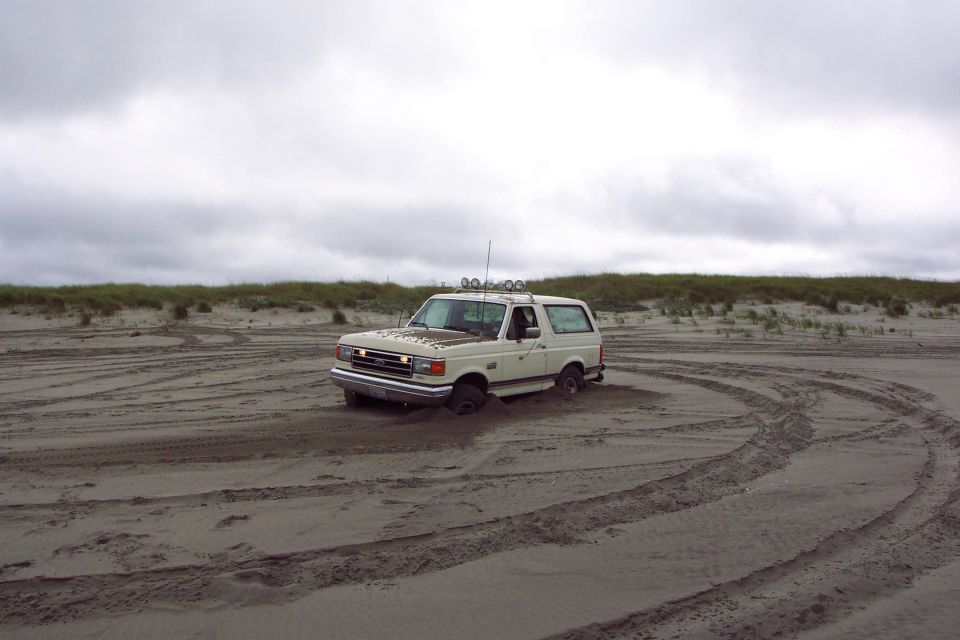 White Ford Bronco SUV stuck in beach sand with tire tracks under cloudy sky