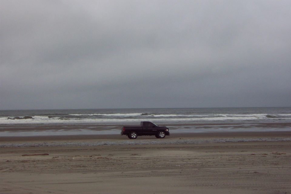 Dark pickup truck driving on a beach under gray skies with waves behind