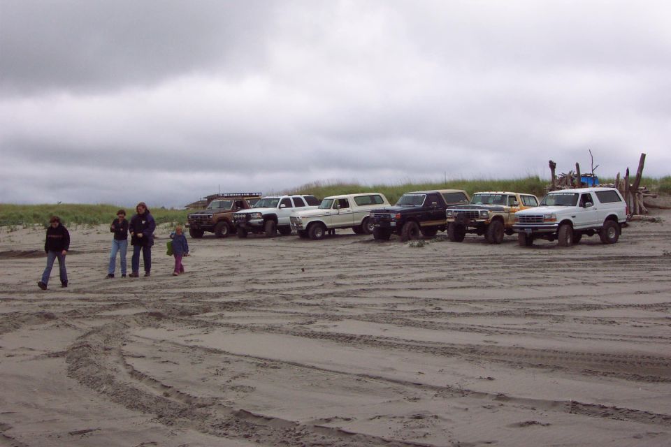 Row of Ford Broncos and trucks parked on an overcast beach with people at left