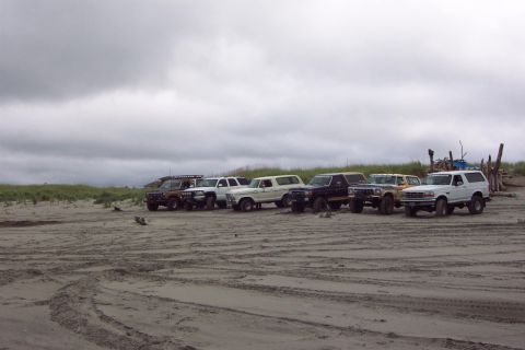 1996 Ford Bronco - Several SUVs and trucks are parked on a sandy beach under a cloudy sky.