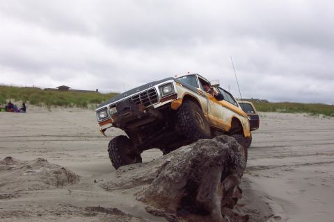 1996 Ford Bronco - This modified Ford Bronco navigates a rocky obstacle on a sandy shore, with another vehicle visible in the background.