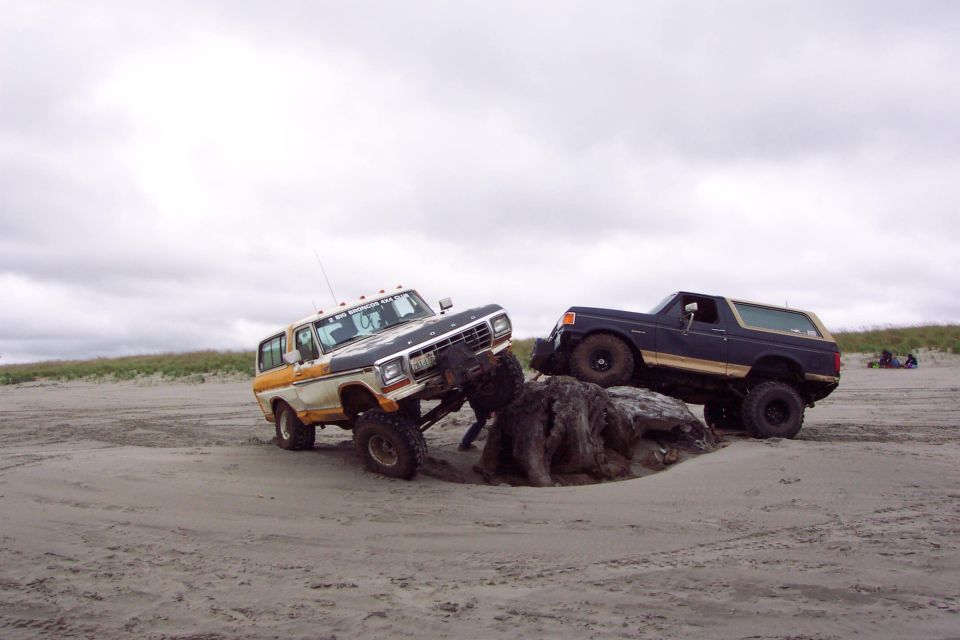 Two lifted Ford Broncos on overcast beach, one tan/white nose-down in sand, one dark blue behind