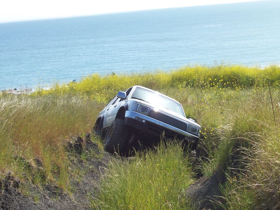 Front three-quarter of a silver Toyota SUV nosed down a grassy ocean-side slope into a rutted dirt trail