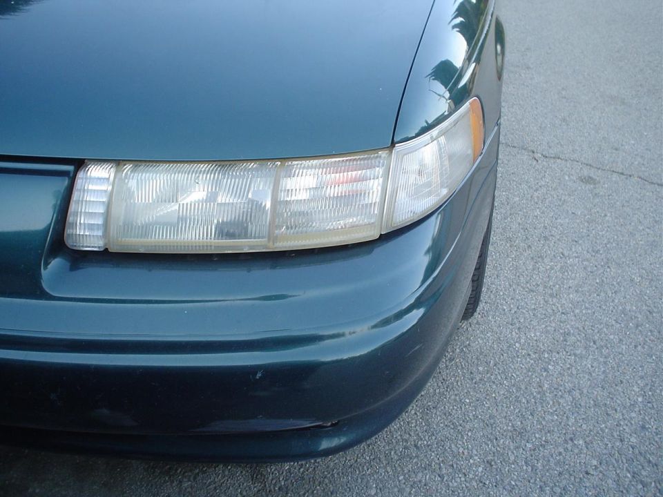 Close-up of a green sedan's left headlight cluster and amber marker over the front bumper