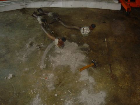 1986 Ford Mustang - Exhaust system components lie on a dusty concrete floor next to a hammer and pry bar.