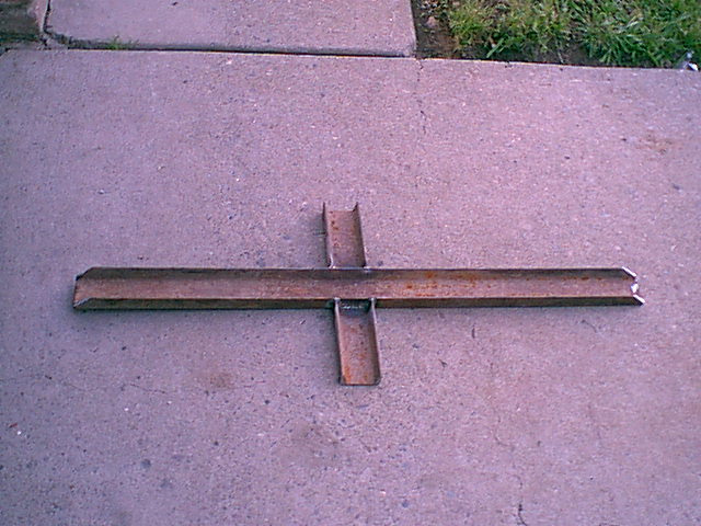 Rusty steel cross-shaped welded metal bracket lying on a concrete driveway