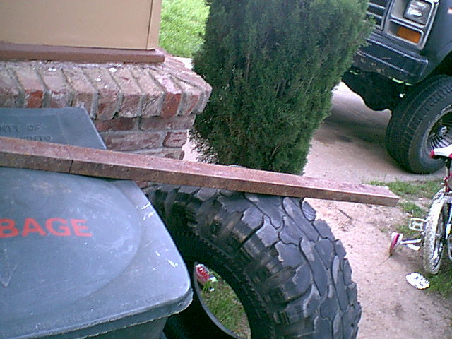 Large mud-terrain tire beside a garbage can with a rusty steel bar on top; dark truck front visible in background