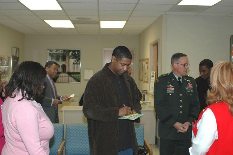 Denzel Washington in a brown coat signs a document in an office with a military officer and others