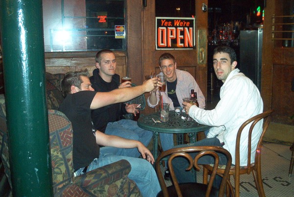 Four men toasting drinks at a bar table under a neon open sign