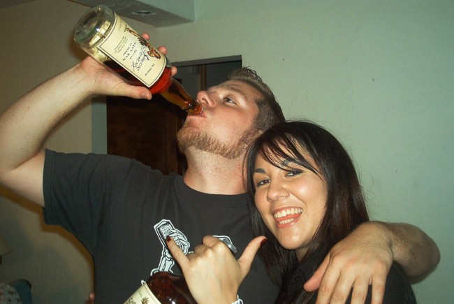 Man drinking from a bottle with his arm around a smiling woman indoors