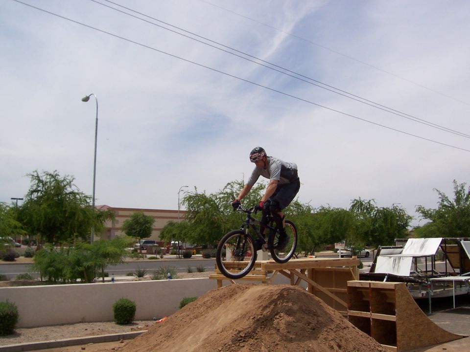 Person on a bicycle mid-air over a dirt ramp, wearing a helmet and casual clothes. A trailer with white coverings is visible to the right