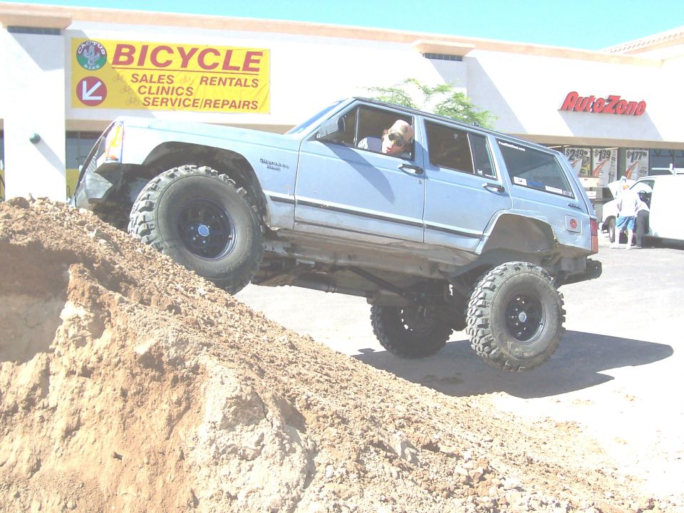 Light blue SUV with large tires climbing a dirt mound, driver visible
