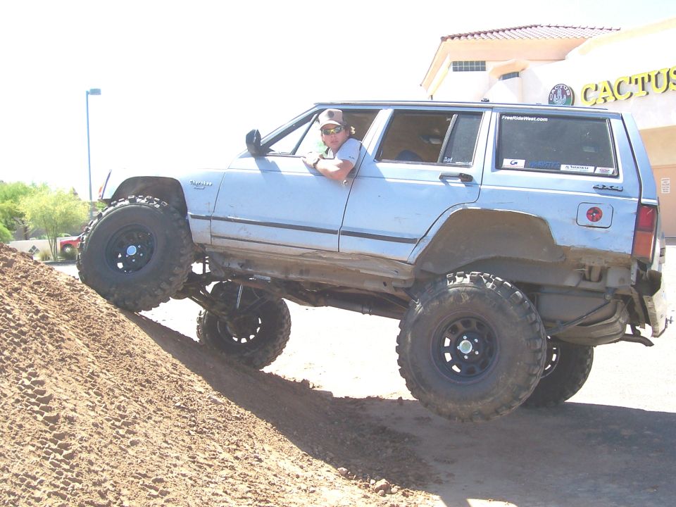 Modified Jeep Cherokee XJ with large tires climbing a dirt mound, driver visible