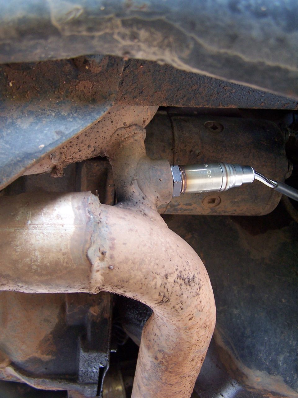 Oxygen sensor screwed into exhaust pipe, with visible rust and dirt on undercarriage components