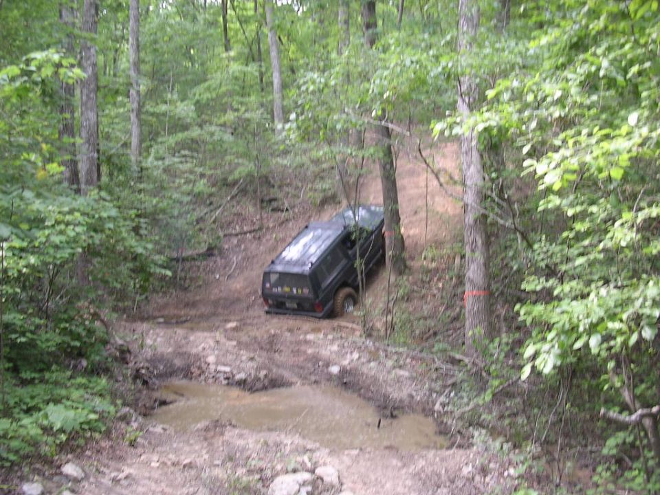 Dark Jeep Cherokee climbing a steep dirt trail in the woods above a muddy creek