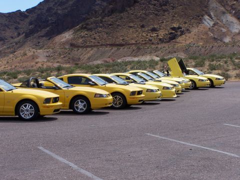 2002 Ford GT - Multiple yellow Ford Mustangs are parked in a row, with one notable example upward-opening scissor doors.