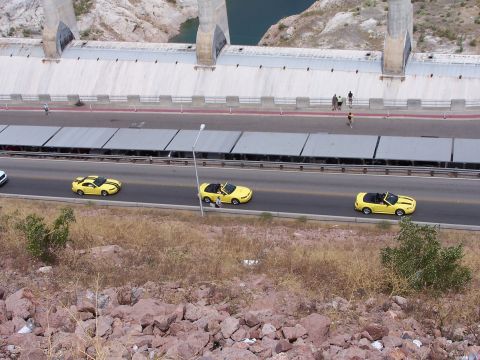 2002 Ford GT - Three yellow convertible sports cars drive on a road near a large dam structure.