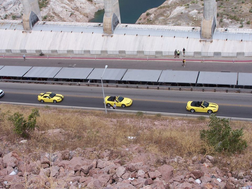 Three yellow convertible sports cars with black stripes drive in a line on a road next to a large dam