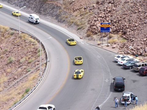 2002 Ford GT - Several yellow convertible sports cars are seen driving along a winding road in Arizona, with a parking lot full of other vehicles nearby.