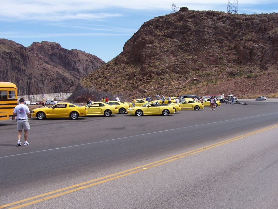 Several yellow Ford Mustangs parked in a lot next to a yellow school bus, with mountains in the background