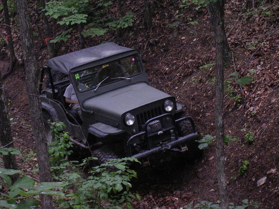 Olive green Jeep CJ-5 with a soft top driving on a steep, wooded dirt trail. Aftermarket front bumper and winch visible