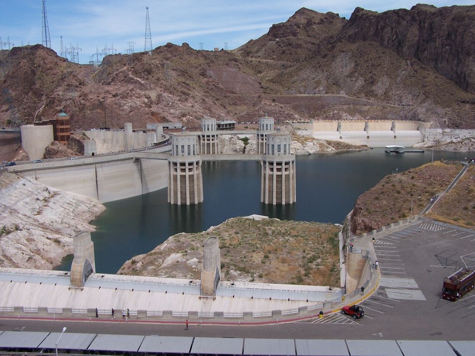 Hoover Dam with water and mountains in background. Two vehicles are visible in the parking lot and on the road