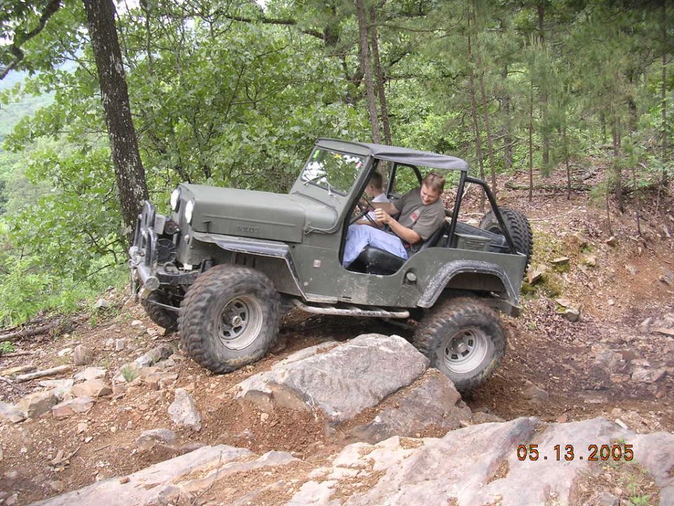 Olive green Jeep CJ-7 off-roading on a rocky trail, with two people inside. The vehicle has large tires and a roll cage