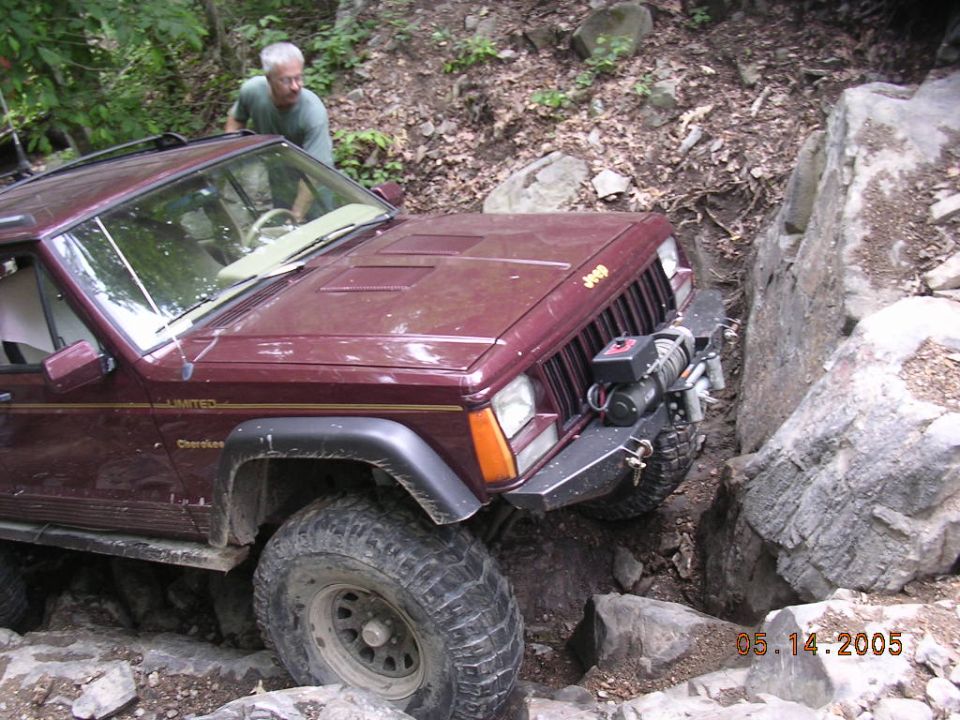 Maroon Jeep Cherokee with winch navigating a rocky trail. Man in green shirt watches